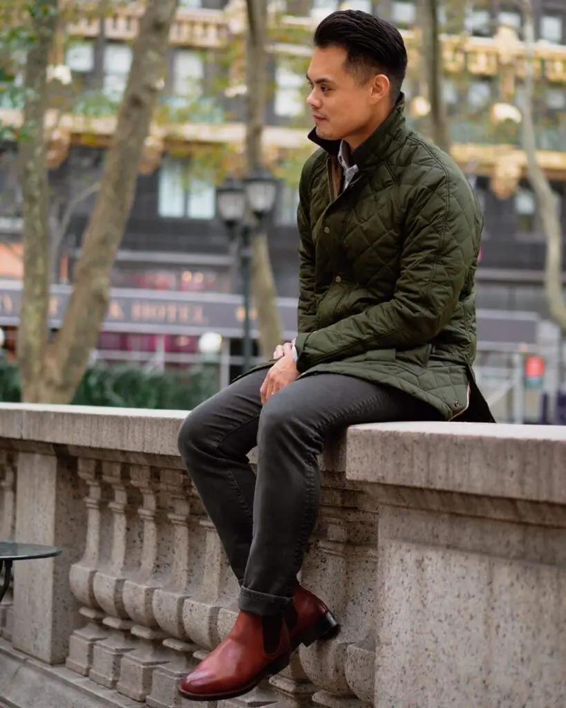 man wearing faded black jeans green quilted jacket and brown chelsea boots sitting on ledge