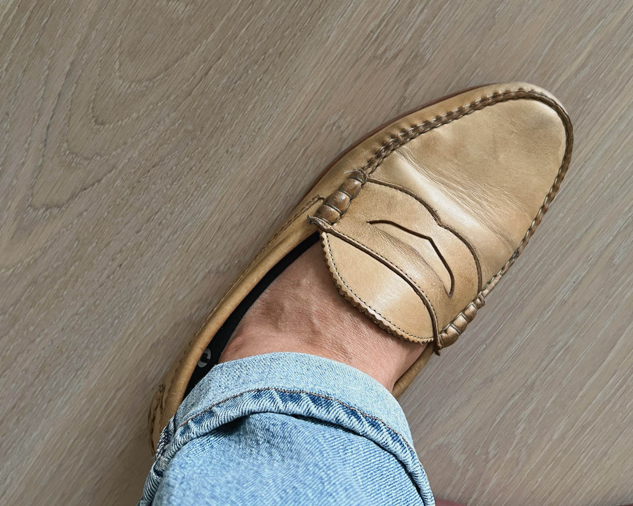 closeup of tan loafer on foot with cuffed light blue jeans and black no show socks on light wood floor