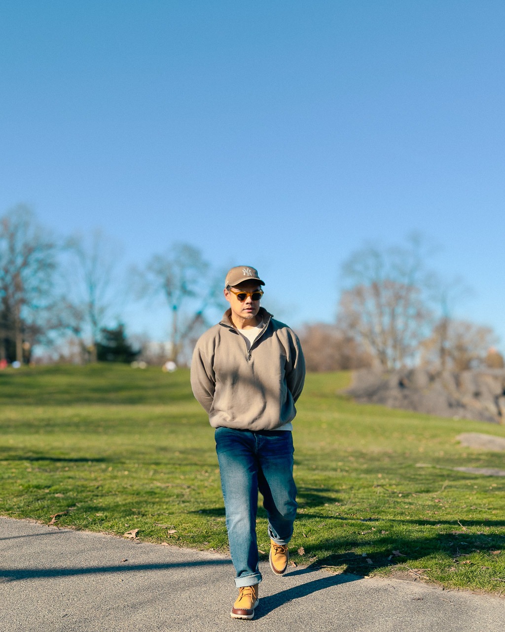 insanely handsome asian man wearing blue jeans quarter zip sweatshirt ballcap and sunglasses in a park with green grass walking towards camera