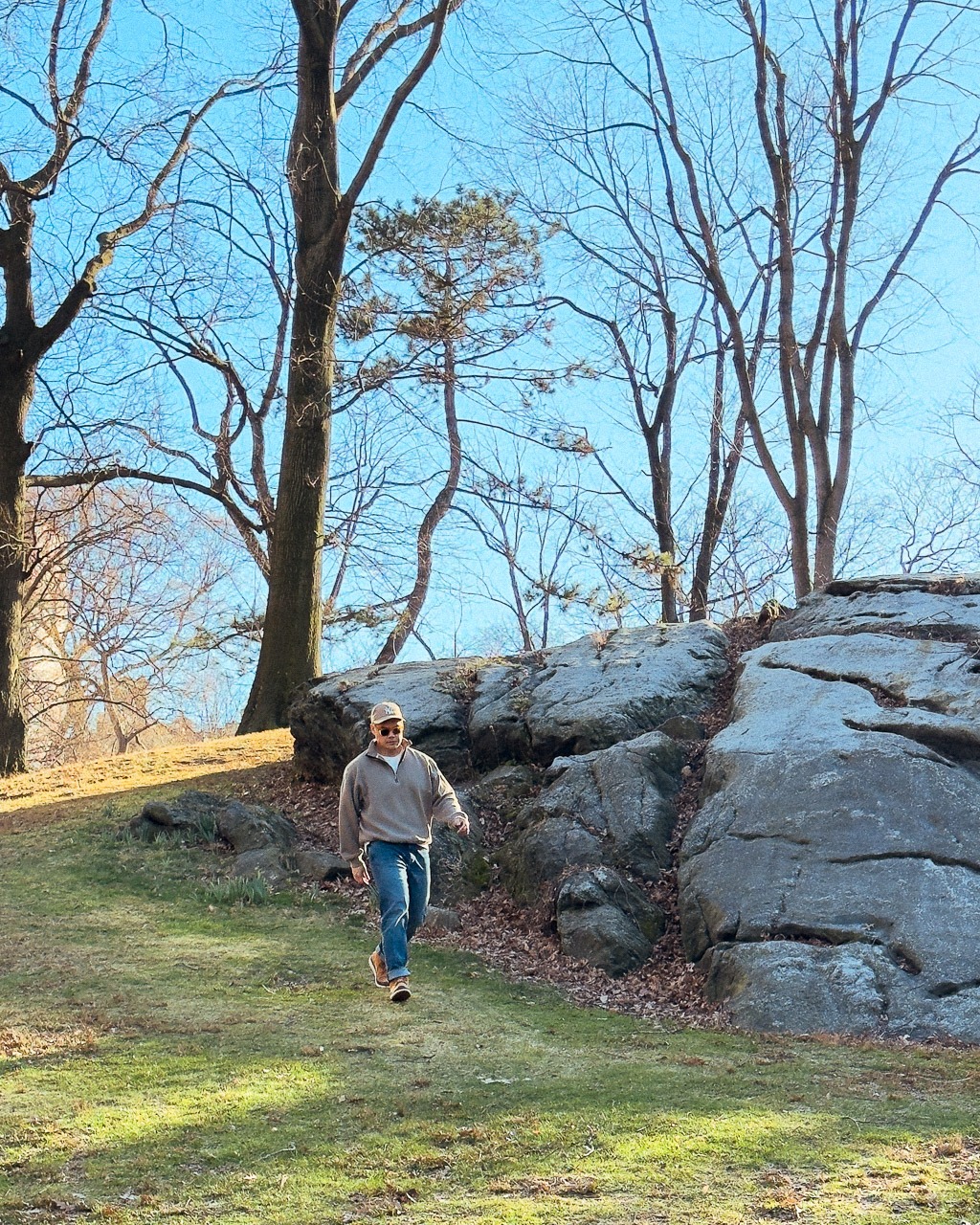 man in jeans and quarter zip sweatshirt and ball cap walking down green grass hill in a park with rock and tall trees in the background